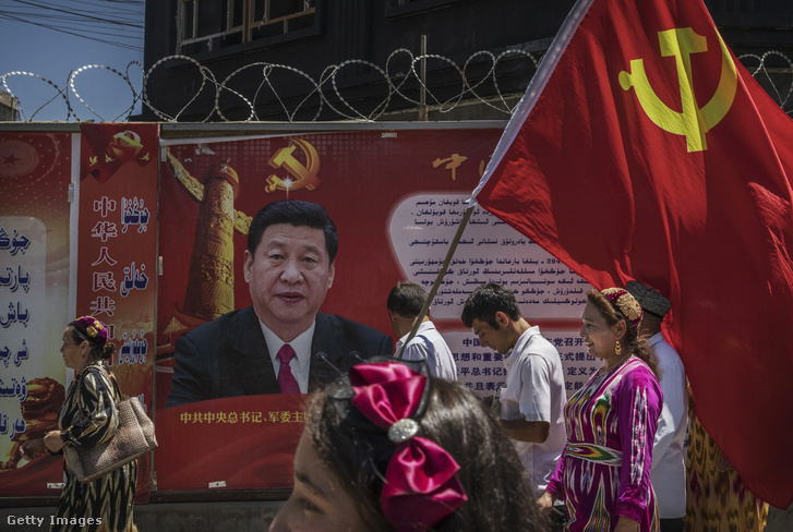 A Kommunista Párt ujgur tagjai zászlóval egy Hszi Csin-ping kínai elnököt ábrázoló plakát előtt 2017. június 30-án a hszincsiangi Kasgar városában.