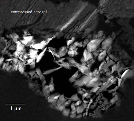Kalcium-alumínium tartalmú szemcse a Wild 2-üstökös porából gyűjtött  54. sz. mintában. A képen a csapdába nagy sebességgel érkező szemcsét lefékező aerogél anyag a sima háttér. Az 1 mikronos méretskála is jelölve van (kép: NASA/JPL)