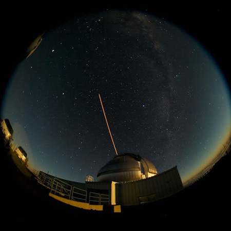 Halszemoptikás felvétel az Északi Gemini távcső mellől, amelyen lézeres műcsillag hasít magasan az égbe.
