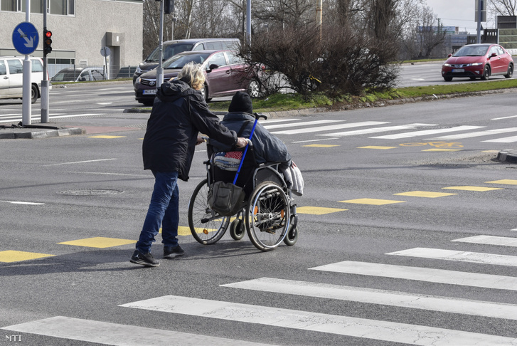 Egy kerekesszékes mozgáskorlátozott személyt tol át kísérője a gyalogátkelőhelyen a IX. kerületben a Könyves Kálmán körúton