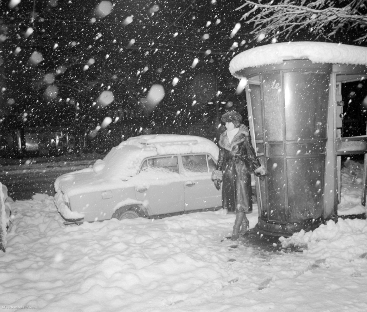 Jellegzetes, gombatetejű műanyag telefonfülkéből lép ki egy járókelő a sűrű havazásban 1980. december elsején. Mellette egy Lada személygépkocsi