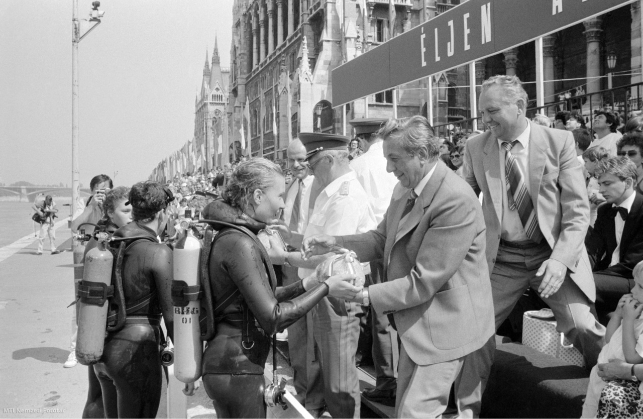 Budapest, 1988. augusztus 20. Könnyűbúvárok új kenyérrel köszöntik a dísztribün vendégeit a budapesti vízi és légi parádén, az alkotmánynapi ünnepségek egyik leglátványosabb attrakciójának keretében. Középen, hátul Fejti György, a Magyar Szocialista Munkáspárt Központi Bizottságának (MSZMP KB) titkára és Kárpáti Ferenc vezérezredes, honvédelmi miniszter (szemüvegben). Kárpáti Ferenc (Putnok, 1926. október 16. – Budapest, 2013. szeptember 27.) hivatásos katonatiszt, honvédelmi miniszter (1985. december 30. – 1990. május 23.). Fejti György (1946–) politikus, 1980. december 3-án lett a KISZ KB első titkára, beosztásából 1984-ben felmentették; 1984. december 7-én az MSZMP Borsod-Abaúj-Zemplén megyei pártbizottság első titkárává választották. 1989. áprilistól a rendszerváltás feltételeiről szóló háromoldalú politikai egyeztető tárgyalásokon az MSZMP tárgyalóküldöttségnek helyettes vezetője volt. 1989. júliusban a Politikai Bizottság helyett felállított Politikai Intéző Bizottságba is bekerült. 1989. októberben visszavonult a politikai élettől, magánvállalkozó lett