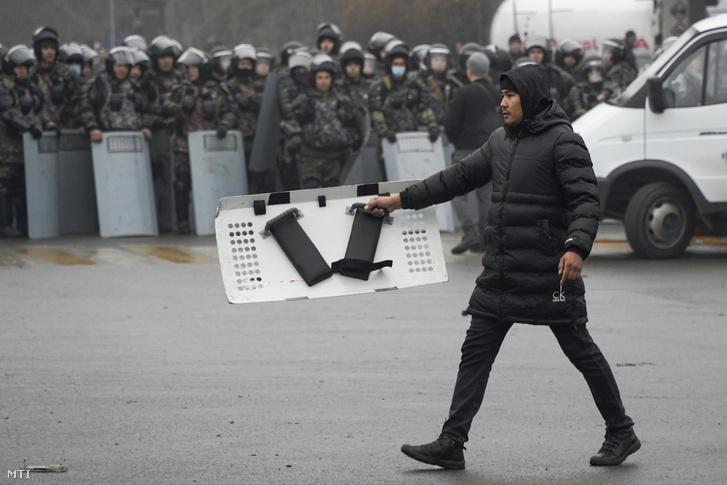Tüntető rendőrségi pajzsot tart maga előtt miközben elhalad a rohamrendőrök sorfala előtt Almatiban 2022. január 5-én