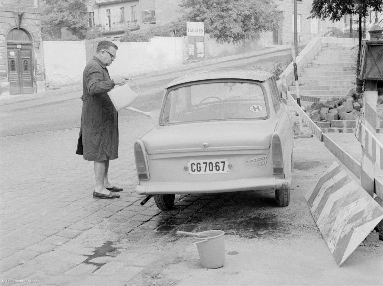 Budapest, July 21, 1969 A man is watering his car in Trabant from a can on the street.  The recording captures the age-old, self-washing car wash - in the foreground is the finished shampoo water and washing brush, and the owner's outfit is a cape suitable for the occasion.  By the way, the date is the date of the American moon landing - which is why the street is quiet and deserted despite Monday.