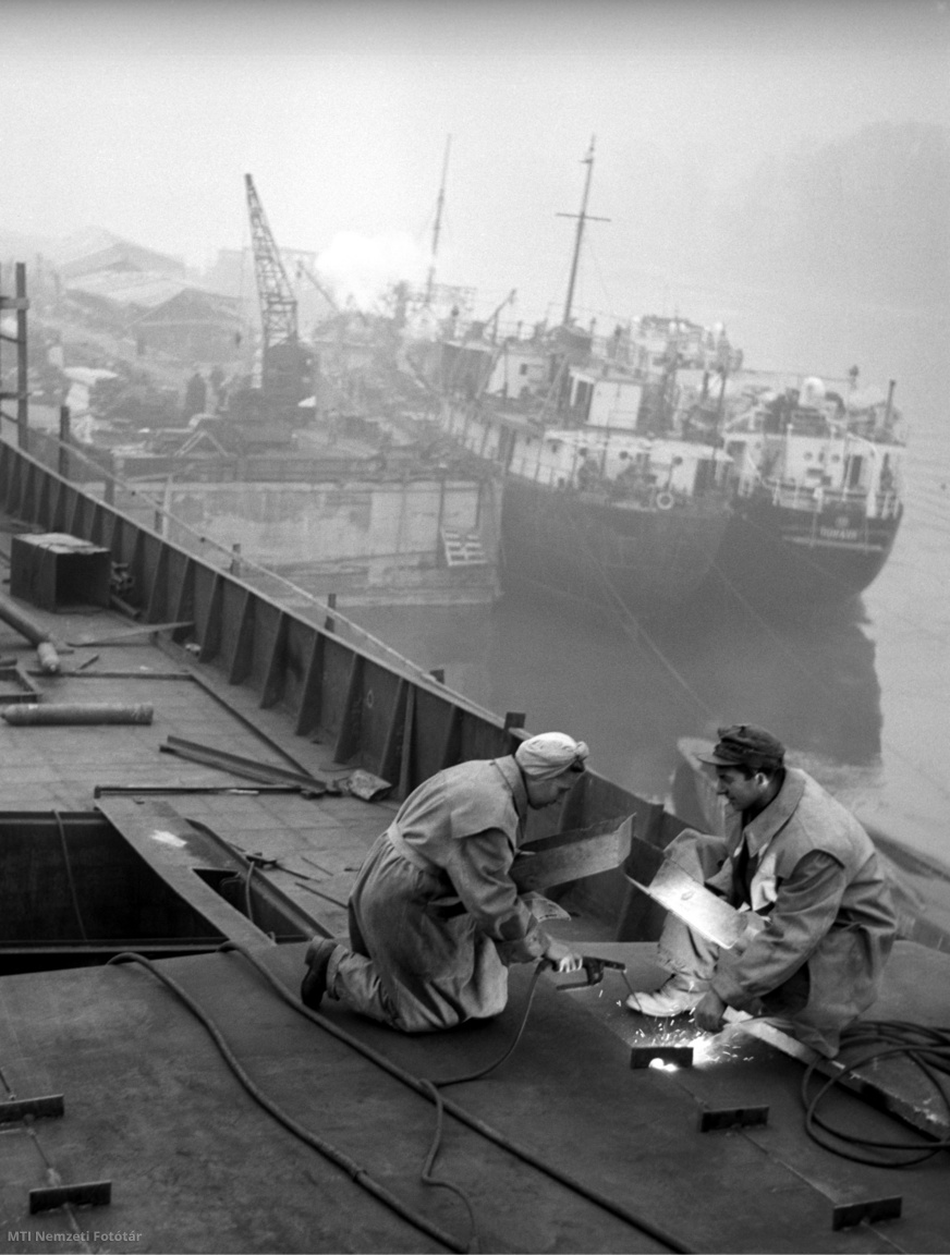 Budapest, 1954. november 2. B. Szilágyi István sztahanovista hajókovács brigádvezető és felesége hegeszt az 56-os számú hajón a Gheorghiu-Dej Hajógyárban rendezett forradalmi műszakon.  Gheorghiu-Dej Hajógyár: a Ganz Danubius Villamossági-, Gép-, Vagon- és Hajógyár-konszernt 1948-ban felosztották, a hajógyár Ganz Hajógyár néven működött tovább. 1951–1962 között Gheorghiu-Dej Hajógyár, majd a Magyar Hajó- és Darugyár Angyalföldi Gyáregysége nevet viselte, 1985-ben Ganz Danubius Hajó- és Darugyár lett
