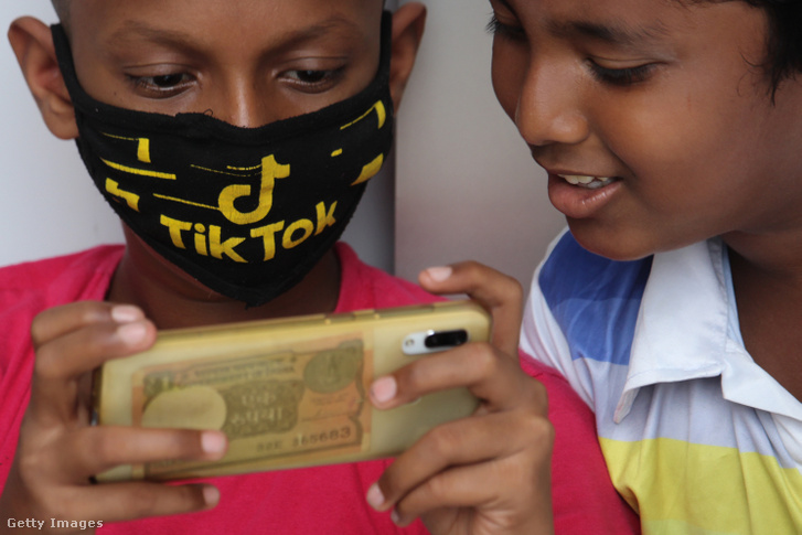 TikTok-logós maszkban telefont néző gyerekek egy koronavírus-járvány miatt bezárt bolt előtt Mumbaiban, Indiában 2020. július 29-én