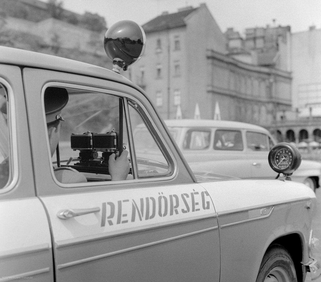 Budapest, 1965. május 18. Rendőrautóba szerelt közúti sebességmérő műszert (traffipax) próbálnak ki a közlekedésrendészet munkatársai a gyorshajtók kiszűrésére a Lánchídnál, a Clark Ádám téren. A Német Szövetségi Köztársaságban (NSZK), a Robot GmbH által gyártott Traffipax márkanevű készülékek nappali és éjjeli körülmények között egyaránt képesek dokumentumszerűen lefényképezni a közlekedési kihágást elkövető gépkocsit. A két felvevőgéppel készült képen látható a jármű rendszáma, valamint az elkövetés napja és időpontja is, gyorshajtás esetén pedig, a sebesség adat. A készüléket egy hónapig kipróbálják, hogy megállapíthassák, mennyire alkalmazható a magyar utakon és ezt követően a szakemberek eldöntik, hogy mennyi berendezést vásároljanak meg. A háttérben a Lánchíd presszó terasza.