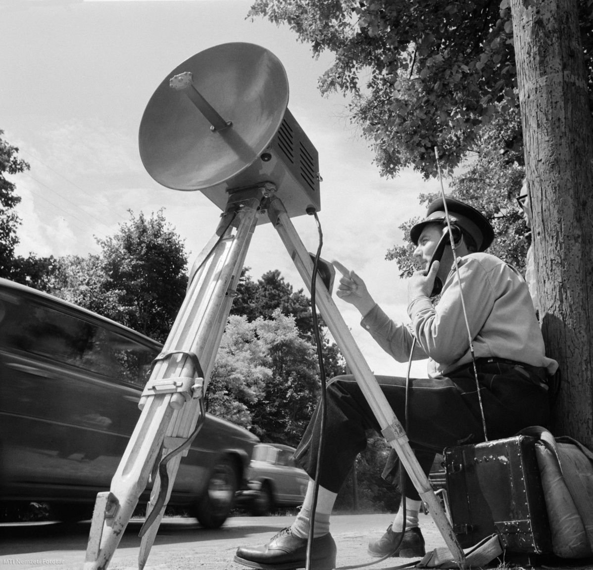 Magyarország, 1965. július 29. Buckó György rendőr törzsőrmester rádiótelefonon jelzi, a közúti sebességmérő műszer (traffipax) segítségével bemért, a sebességkorlátozást túllépő gépkocsi rendszámát az úton távolabb várakozó kollégáinak, hogy azok intézkedhessenek. A felvétel készítésének pontos helye ismeretlen.