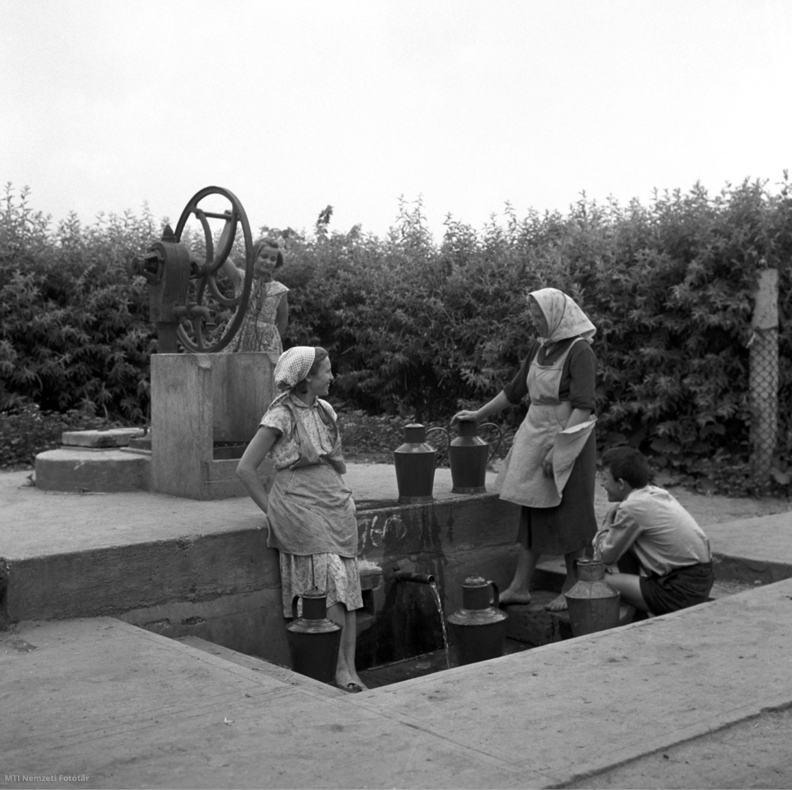 Magyarország, 1960. június 20. Asszonyok beszélgetnek, mellettük kanna, háttérben fiatal lány vizet húz a kerekes kútból