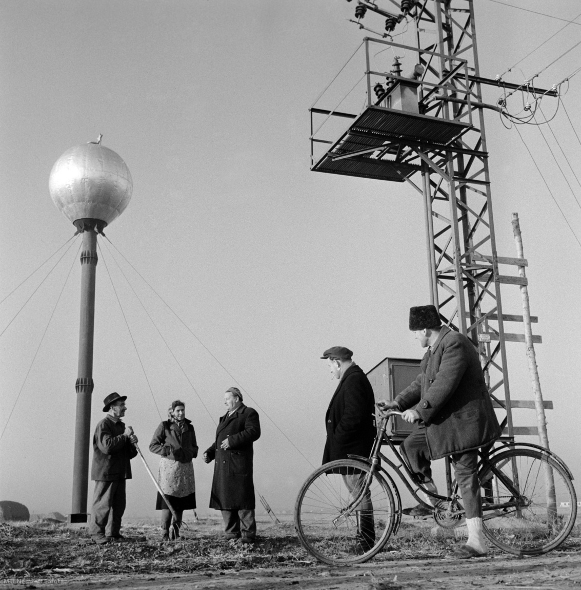 Magyarország, 1959. november 27. Termelőszövetkezeti (tsz-) tagok beszélgetnek munka közben a gazdaság földjén. Háttérben jobbra egy villanyoszlopra szerelt transzformátor, balra egy víztorony (hidroglóbusz). A felvétel készítésének helyszíne ismeretlen.