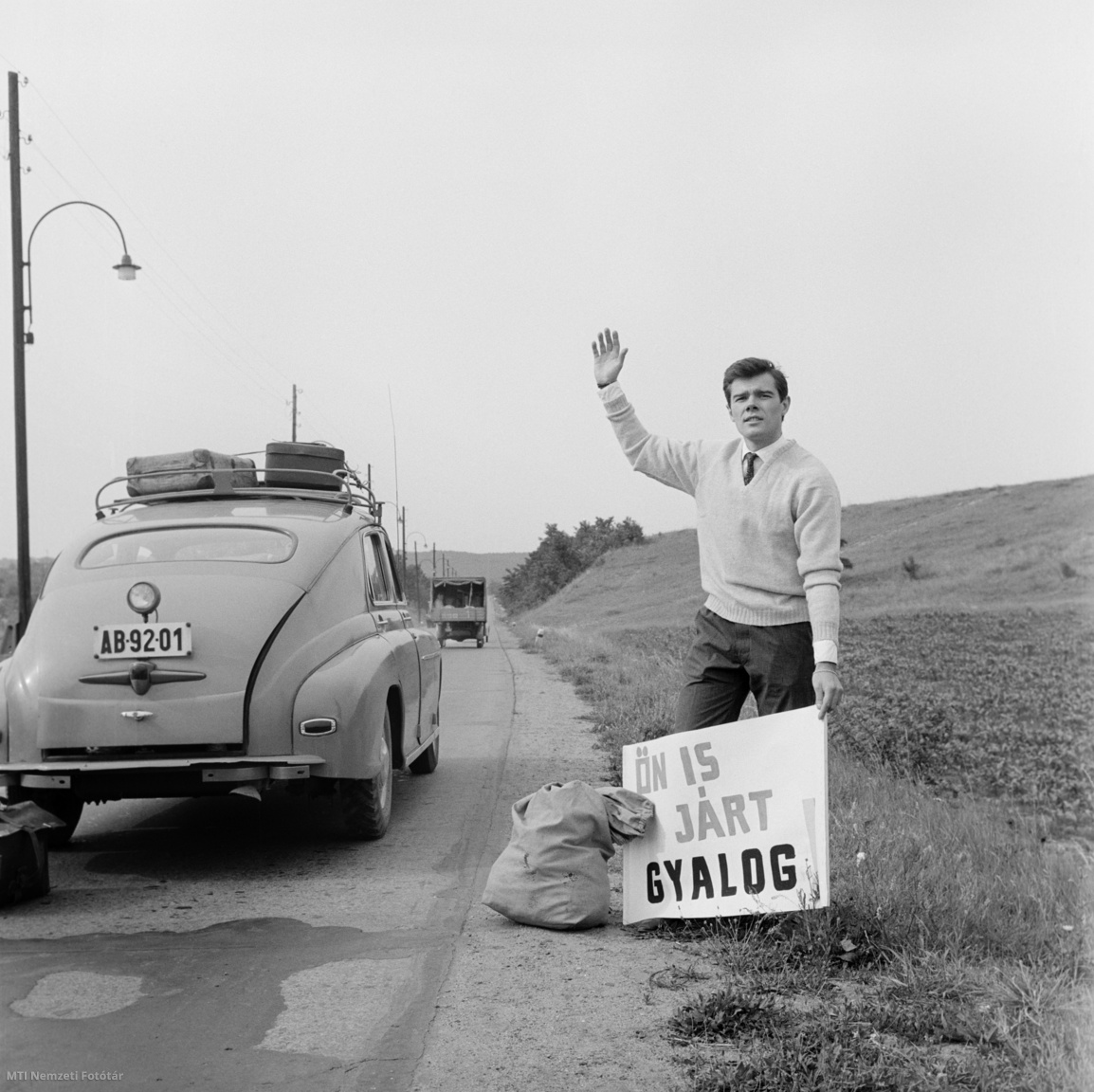 Magyarország, 1962. július 1. Tovább utazásához autókat próbál lestoppolni egy férfi az országút széléről, egy Ön is járt gyalog feliratú tábla segítségével. A felvétel készítésének pontos helye ismeretlen.