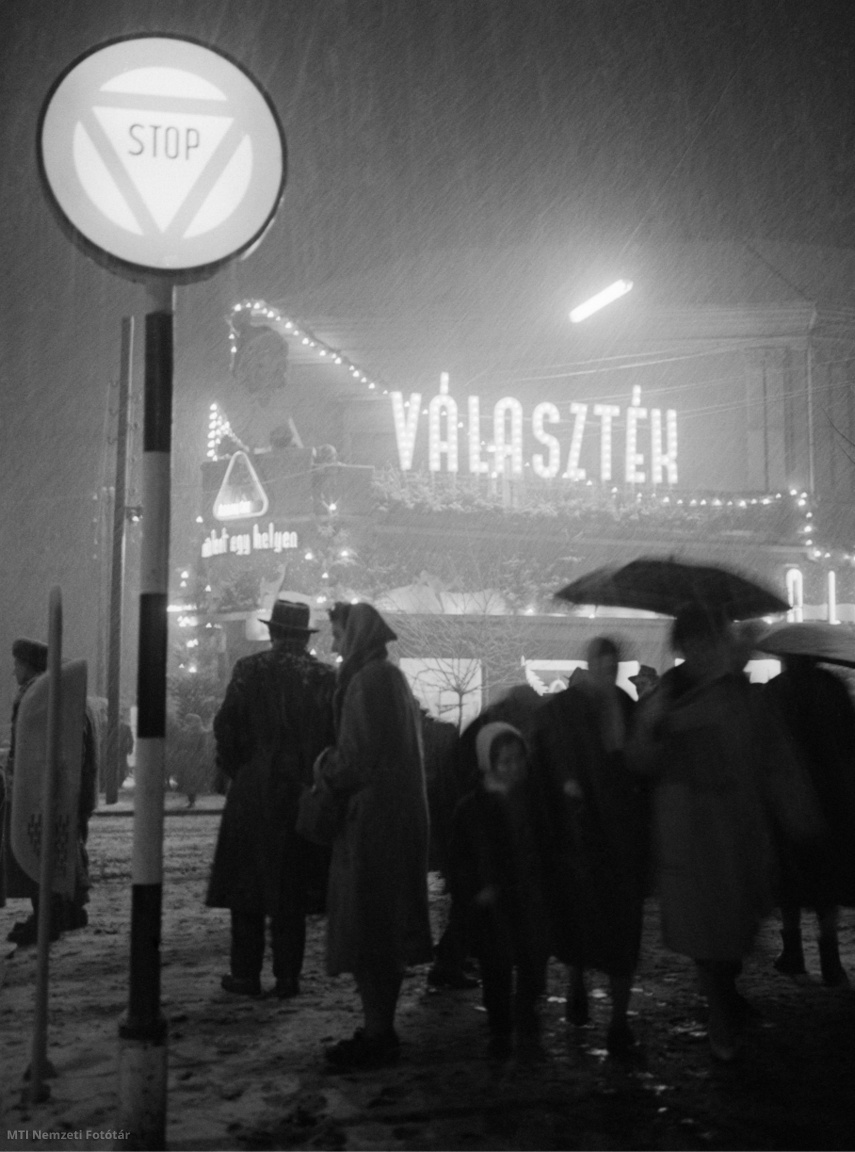 Magyarország, 1961. december 14. Járókelők a téli havazásban a karácsonyi vásárt hirdető neonreklámok alatt egy város utcáján. A felvétel készítésének pontos helye ismeretlen