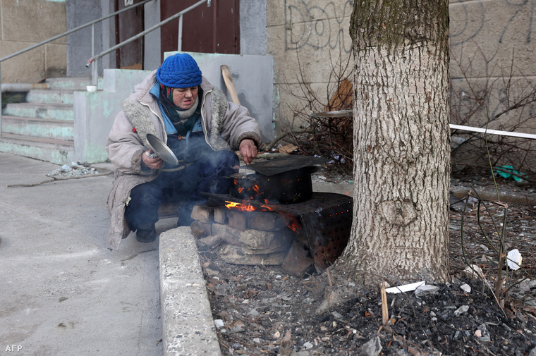 Egy idős asszony főz egy társasház bejárata mellett a szabadban Bahmutban, 2023