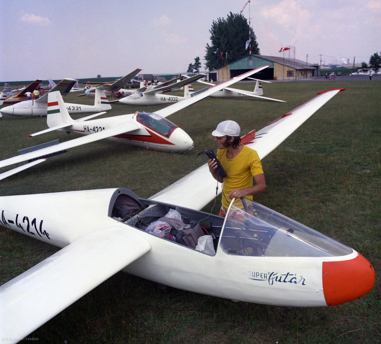 1976. július 4. Folyik a rádiók ellenőrzése a felszállásra váró vitorlázó repülőgépeken a dunaújvárosi (Kisapostag) MHSZ (Magyar Honvédelmi Szövetség) sportrepülőtéren az országos válogató bajnoksága mezőnyében. Az előtérben egy Rubik R-22S Super Futár típusú vitorlázó repülőgép, mögötte egy lengyel gyártású SZD-12 Mucha.