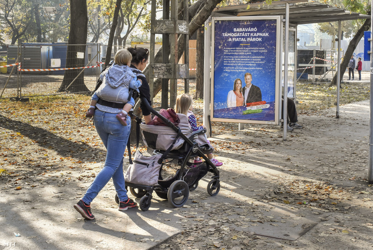 Egy fiatal anya gyermekeivel sétál egy babaváró támogatást hirdető plakát mellett a Városligetben