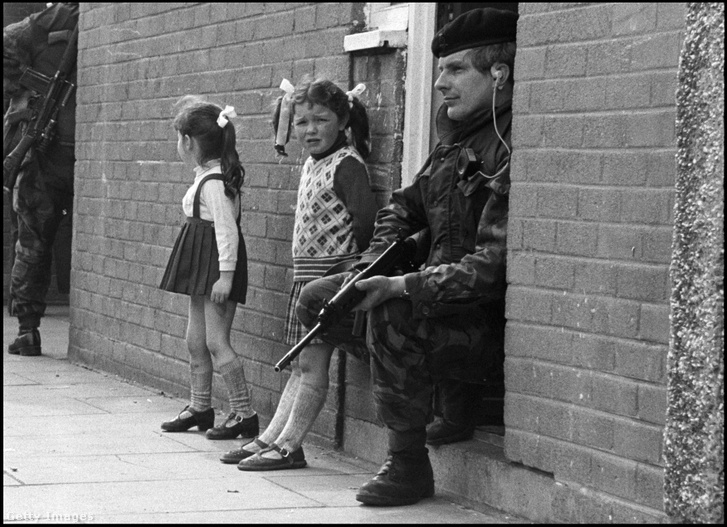 Kislányok állnak az utcán, miközben egy brit katona ül az otthonuk ajtajában a hadsereg hadművelete során Belfast New Lodge Road nevű republikánus kerületében, 1976. április 19-én