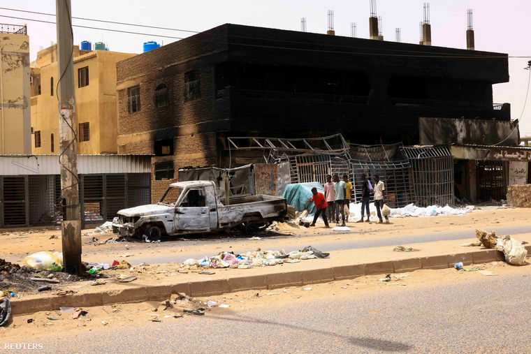 Helyiek sétálnak egy kiégett autó és házak mellett a szudáni főváros, Kartúm központi piacánál, 2023