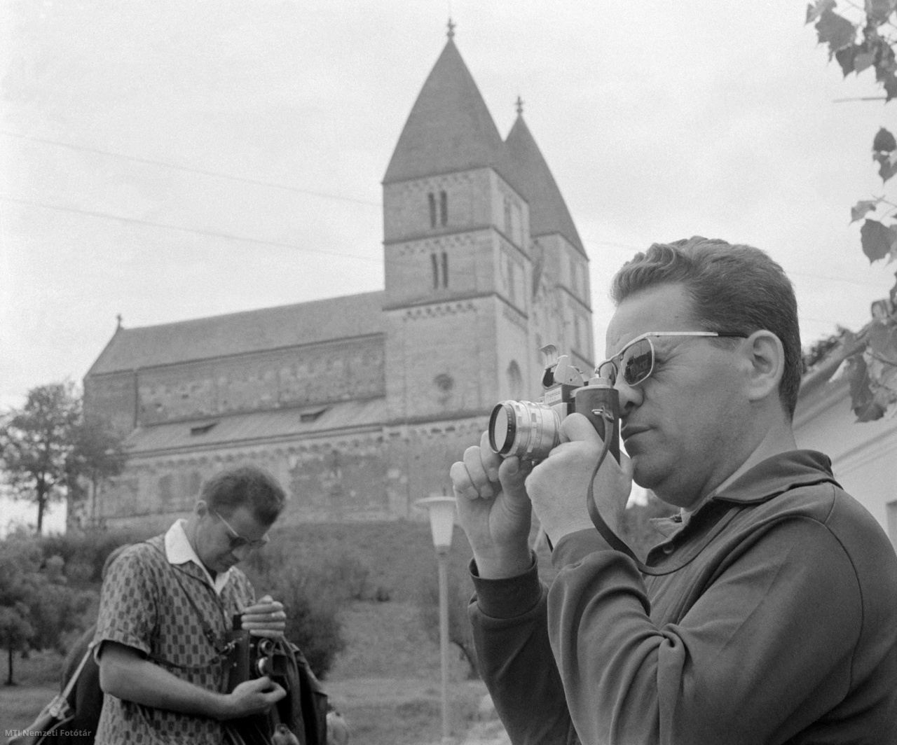 Ják, 1965. július 17. Egy férfi fényképez a VEB Kamera-Werke Drezda Niedersedlitz fényképezőgépgyár Praktica márkanevű, tükörreflexes, kisfilmes fényképezőgépével a jáki templom, az egykori jáki bencés apátság monumentális bazilikája előtt. A Népművelési Intézet az idén Szombathelyen rendezte meg a hagyományossá vált nyári fotós szakmai továbbképző tanfolyamot, amelyen az országos fotóamatőr-mozgalom vezetői, fotóművészek vesznek részt. A 80 fotográfus a tíznapos tábor idején esztétikai, képzőművészeti előadásokat hallgat, kiállításokat rendez és vitat meg, valamint fotótúrát tesznek Jákra és Kőszegre