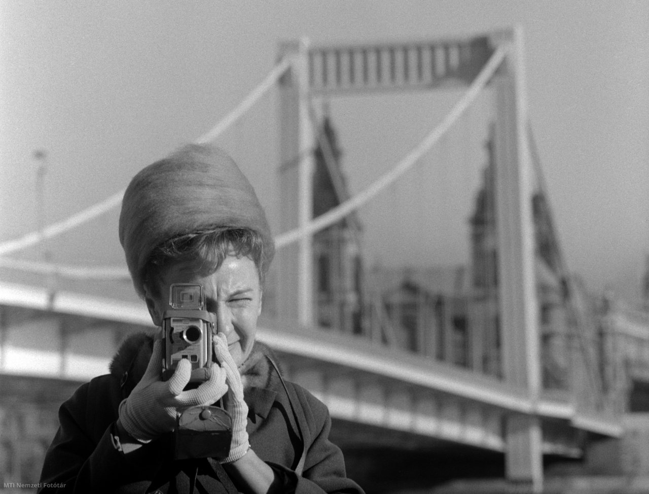 Budapest, 1965. december 1. Egy filmfelvevőgéppel felvételt készítő nő a decemberi napsütésben az Erzsébet híd budai hídfőjénél. A háttérben a Budapest-Belvárosi Nagyboldogasszony Főplébánia-templom (Belvárosi plébániatemplom)