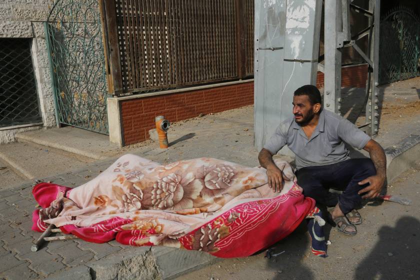Egy férfi a bombázások egyik áldozata mellett ül egy romos utcán Gázában.