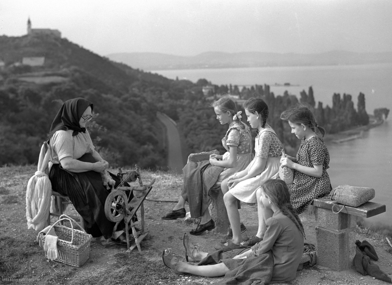 Tihany, 1954. június 22. Padon, földön ülő leányok nézik a rokkázó asszonyt, mögöttük a Tihanyi Bencés Apátság