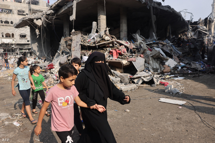 Egy nő és gyerekek sétálnak el a gázai Al-Shatee táborban 2023. október 28-án az izraeli bombázások során lerombolt ház mellett