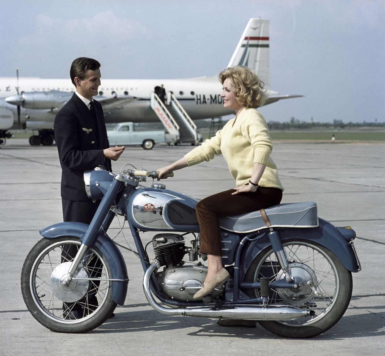 Budapest, 1963. május 10. Modell a Csepeli Motorkerékpárgyár egyik új termékén, egy 250 köbcentiméter hengerűrtartalmú motorral szerelt, Pannónia típusú motorkerékpáron ülve beszélget egy pilótával a Ferihegyi repülőtér betonján a reklámcélra készült felvételen. A háttérben a Magyar Légiforgalmi Vállalat (MALÉV) HA-MOA lajstromjelű légcsavaros IL–18V típusú repülőgépe