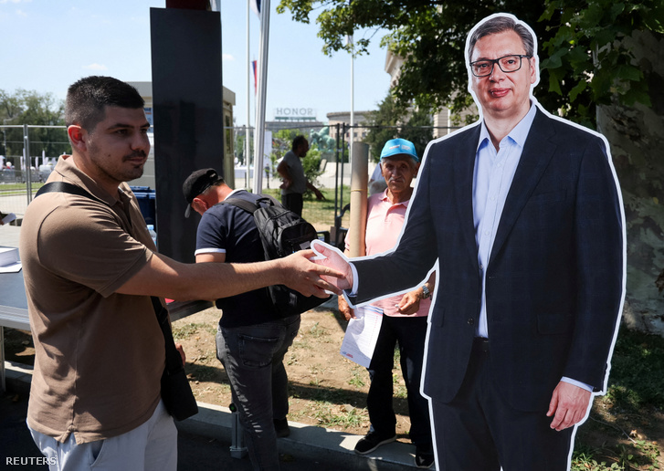 Egy férfi a szerb elnököt, Aleksandar Vucicot ábrázoló kartonfiguras kezét fogja, miközben a kormányzó Szerb Haladó Párt (SNS) támogatói gyűlésre gyülekeznek Belgrádban, 2026. június 27-én