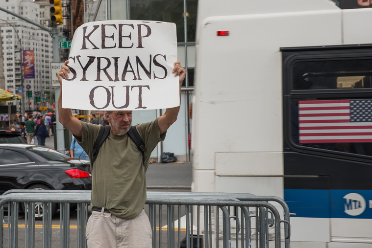 Egy magányos tüntető menekültellenes plakáttal áll egy menekültpárti demonstráció mellett.